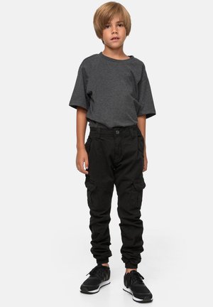 Ung kille står, iklädd en mörkgrå t-shirt, svarta cargobyxor och svarta sneakers mot en ljus, enfärgad bakgrund.