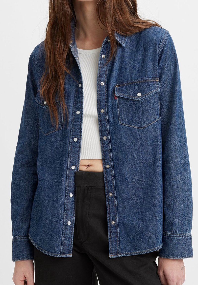 Femme portant une chemise en jean bleue ouverte à boutons par-dessus un crop top blanc côtelé et un pantalon noir, avec de longs cheveux bruns visibles.