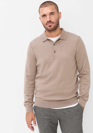 Uomo con capelli corti e barba, che indossa un maglione polo beige a maniche lunghe e pantaloni grigi, in piedi davanti a uno sfondo bianco semplice.