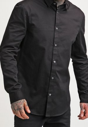 Schwarzes Hemd mit langen Ärmeln und Knöpfen, getragen von einer Person mit tätowierten Händen, stehend vor einem schlichten, hellen Hintergrund.