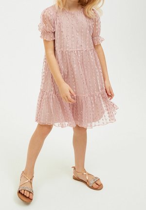 Robe rose en tissu transparent, avec des manches bouffantes, des couches plissées et des embellissements à pois. Portée avec des sandales en bronze à lanières.