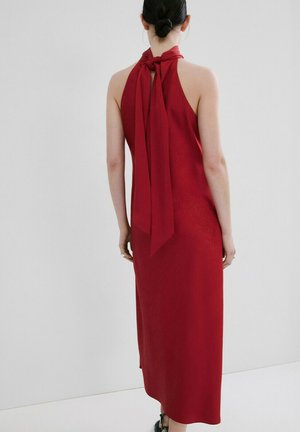 Femme aux cheveux foncés attachés en chignon, portant une robe rouge sans manches à col halter nouée au cou, debout devant un fond clair uni.