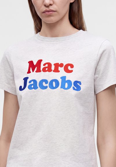 Γυναίκα φοράει ένα ανοιχτού γκρι T-shirt με το "Marc" με κόκκινα και το "Jacobs" με μπλε τυπωμένα στο μπροστινό μέρος.