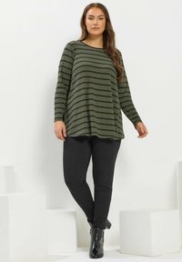 Nainen, joka pitää yllään oliivinvihreää pitkähiharaista raidallista paitaa, mustia slim-fit housuja ja mustia nilkkuremmitossuja, seisoo valkoista taustaa vasten.