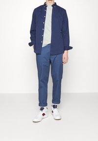 Merivoimien nappipaita vaaleanharmaan t-paidan päällä, yhdistetty sinisiin kapeisiin housuihin ja valkoisiin lenkkareihin, joissa on punaisia ja sinisiä yksityiskohtia.