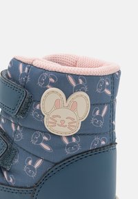 Blåa och rosa barnstövlar med kanintryck, kardborreband och ett kaninansiktsmärke på sidan. Mjuk tygkrage och texturerat material.