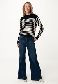 Pullover a righe nere e crema con colletto rotondo, abbinato a jeans a gamba larga blu scuro e tacchi bianchi, su uno sfondo neutro.