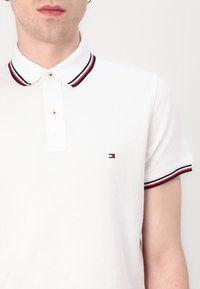 Polo blanc avec une finition texturée, doté d'une patte de boutonnage à deux boutons, d'un col rayé rouge et marine, et d'un petit logo de marque sur la poitrine.