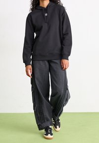 Felpa nera con cappuccio, tasca frontale e coulisse, abbinata a pantaloni cargo grigi oversize con tasche laterali e strisce a contrasto. Sneakers nere con suola chiara.