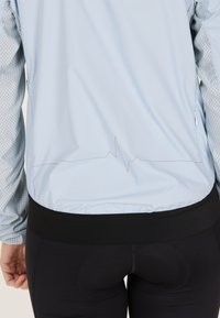 Helles blaues Jackett mit Netzärmeln, das ein Herzschlaglinienmuster entlang des Rückens aufweist. Schwarzer Bund, kombiniert mit schwarzen Leggings.