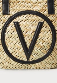 Bolso de tote de paja trenzada con un gran logo en forma de "V" de cuero negro dentro de un acento circular. Cuenta con asas de cuero negro y una textura natural.