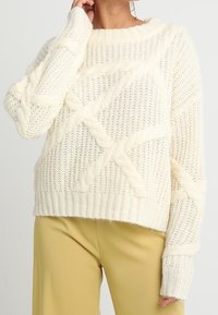 Krämfärgad kabelstickad tröja med texturerat mönster, bred ribbad fåll och långa ärmar. Mjuk, kraftig material med en avslappnad passform.