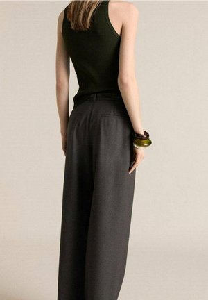 Kobieta widziana od tyłu, ubrana w ciemnozielony top bez rękawów, szerokie ciemnoszare spodnie i liczne bransoletki na prawej ręce.