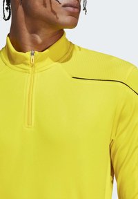 Un pull de sport jaune avec un col haut, une fermeture éclair mi-longue, des accents texturés aux épaules et des coutures noires contrastantes le long des coutures.