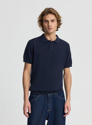 Giovane uomo con capelli biondi ondulati che indossa una polo blu navy a maniche corte e jeans blu scuro, in piedi davanti a uno sfondo semplice.