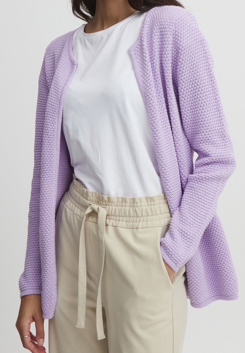 Lavendelstickad cardigan med en strukturerad mönster, öppen fram, långa ärmar och en avslappnad passform, kombinerad med en vit t-shirt och beige byxor.