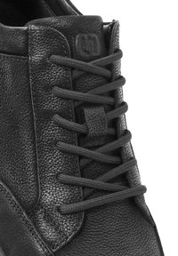 Zapato de cuero negro con superficie texturizada, que presenta cordones gris oscuro y una etiqueta de marca cerca del tobillo. Primer plano de la sección superior.