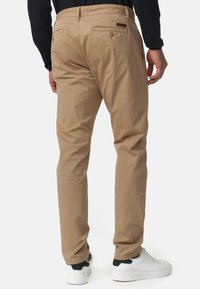Pantalons en coton beige au design ajusté, dotés de deux poches arrière et d'une taille élastique. Portés avec des baskets blanches.