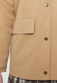 Μπεζ μπουφάν με ομαλή υφή, που διαθέτει τσέπη στο στήθος και στρογγυλά κουμπιά από ορείχαλκο μπροστά. Ορατό καρό σχέδιο από κάτω.