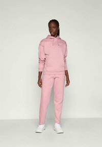 Pink sweatshirt and matching pants in soft cotton blend. Features a hood, front pocket, and subtle logo detailing. Worn with white shoes.