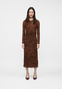 Pruun pikkade varrukatega kleit, millel on südamekujuline must leopardi muster, koos kehakohase vöökohtade ja voolava midi-pikkuse siluetiga. Kombineeritud burgundi tooni kingadega.