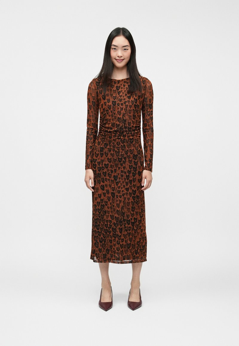 Pruun pikkade varrukatega kleit, millel on südamekujuline must leopardi muster, koos kehakohase vöökohtade ja voolava midi-pikkuse siluetiga. Kombineeritud burgundi tooni kingadega.