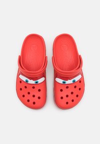 Red clogs with cartoon eyes and eyebrows. Made of smooth plastic, featuring ventilation holes, back strap, and a textured footbed.