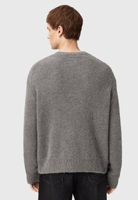Giovane uomo di spalle che indossa un maglione grigio largo e jeans neri, su uno sfondo grigio chiaro uniforme.