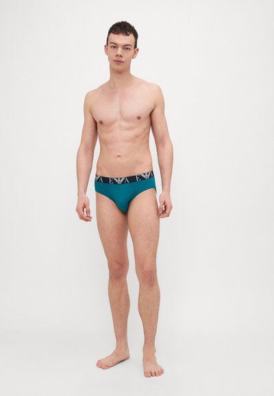 Mutande da uomo color teal con cintura nera che presenta un logo bianco. Il tessuto appare liscio, con una vestibilità aderente e un design a copertura minima.