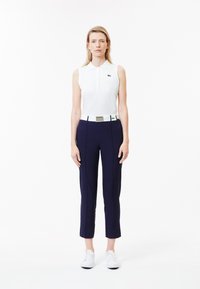 Witte mouwloze polo met kraag, marineblauwe tapered broek en witte schoenen. Inclusief groene logo-accenten en een zilveren riem.