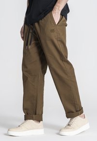 Bruine, relaxte broek met een elastische tailleband en trekkoord, zijzakken en een subtiele geborduurde logo. Gemaakt met lichtbeige sneakers.