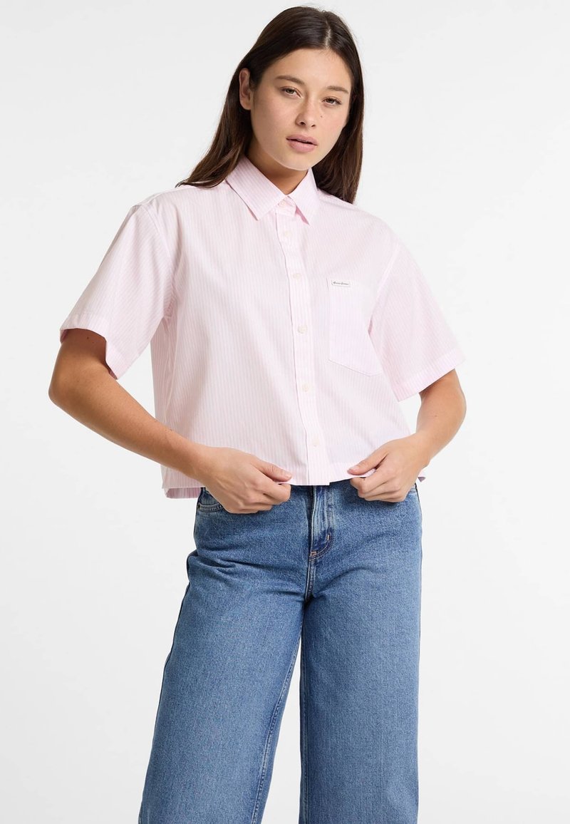 Camicia rosa chiaro a maniche corte con colletto, decorata con righe verticali e una tasca sul petto, indossata con jeans blu a vita alta.