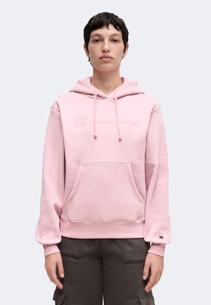 Person iført en lyserød Champion-hoodie med frontlomme og snore, der står mod en ensfarvet lys baggrund.