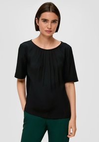Zwarte korte mouwen top met een ronde halslijn en geplisseerde details. Glad materiaal met een lichte glans. Gemaakt met groene broeken.