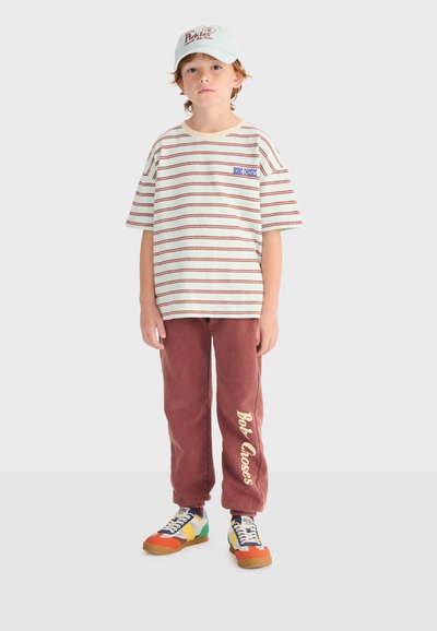Boy wearing a white cap, cream and red striped t-shirt, maroon sweatpants with text, and colorful sneakers, standing facing forward.