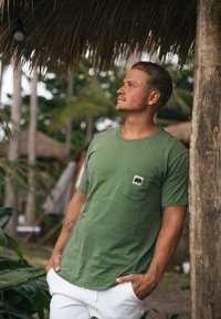 Zelené bavlněné tričko s kulatým výstřihem a kapsičkou na prsou, na které je černé logo slona. Obléknuté s bílými šortkami v přírodním prostředí.