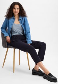 Een denim overhemd over een lichtblauwe tanktop, gecombineerd met marineblauwe broeken en zwarte instapschoenen met dikke zolen, zittend op een stoel.