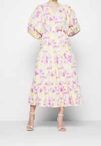 Abito floreale con scollatura a V, maniche a sbuffo e vita cintrata. Tessuto giallo con motivi rosa e viola chiaro, gonna a strati, abbinato a tacchi bianchi.