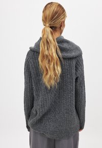 Szary sweter z dzianiny z szerokim kołnierzem i prążkowaną fakturą, o luźnym kroju i obniżonych ramionach. Tylny widok ukazuje subtelne opadanie. 