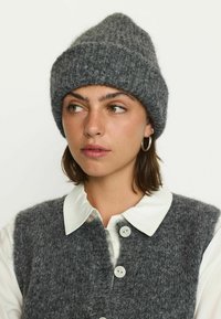 Young woman wearing a chunky gray knit hat and matching buttoned vest over a white collared shirt, looking to the left.