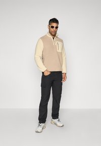 Beige og kremfleece genser med brystlomme, kombinert med svarte cargo bukser og grå treningssko. Tilbehøret inkluderer hvite solbriller.