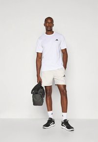 Biała koszulka Adidas, jasnoszare szorty z logo, czarne buty, białe skarpetki, trzyma czarną torbę sportową. Prosty, sportowy design.