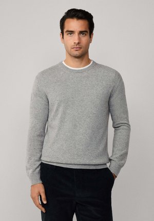 Man in een lichtgrijze ronde hals trui en zwarte broek, staande met één hand in de zak tegen een effen lichte achtergrond.