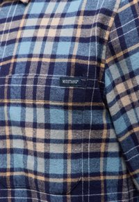 Blauw en beige geruite flanellen shirt met een zak, met het merklabel "MUSTANG®" op de voorkant genaaid. Geweven katoenen stof.