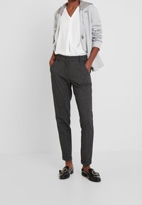 Šedé kalhoty z vlněné směsi s upraveným střihem, vybavené předními kapsami a zapínáním na knoflík, v kombinaci s bílým tričkem s výstřihem do "V" a šedou mikinou s kapucí.