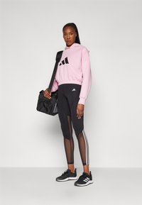 Rožinė trumpa palaidinė su juodu logotipu, derinama su juodomis leggingais su tinkleliu ir baltomis juostelėmis, bei juodais sportiniais batais. Juoda rankinė pakabinta ant peties.