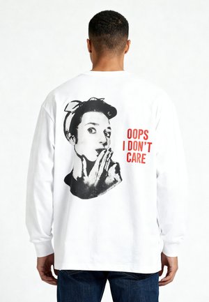 Homme portant un t-shirt blanc à manches longues avec un graphisme rétro en noir et blanc représentant une femme et un texte rouge « OOPS I DON’T CARE » dans le dos.