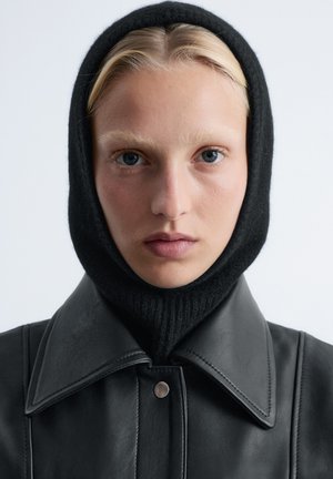 Schwarze Lederjacke mit großem Kragen und silbernen Druckknöpfen. Das Modell trägt eine schwarze gestrickte Sturmhaube. Glatte Textur, minimalistisches Design.