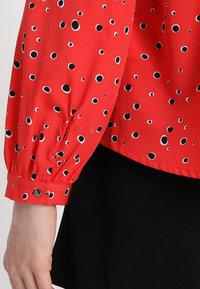 Czerwona bluzka w czarne i białe kropki. Posiada bufiasty rękaw, mankiet zapinany na guziki oraz gładką teksturę materiału.