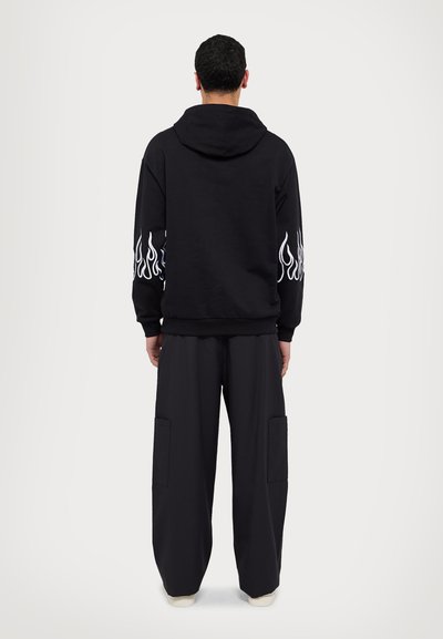 Sudadera negra con capucha y diseños de llamas en blanco en las mangas, combinada con pantalones negros holgados que tienen grandes bolsillos laterales.